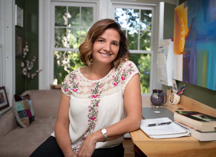 A woman with shoulder-length brown hair is smiling while sitting at a wooden desk with a laptop and notebooks in a well-lit room with large windows.